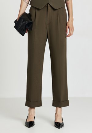 Pantalons taillés vert olive avec des plis, ourlets retroussés et une coupe droite, associés à des escarpins noirs à bout pointu et à une pochette noire.