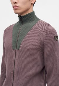 Homme portant un pull en laine zippé avec un col montant vert et un corps texturé mauve, montrant une petite pièce sur le haut du bras.