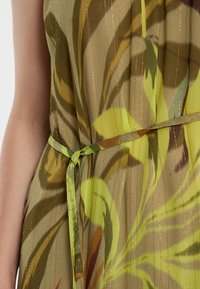 Robe sans manches avec un motif de feuillage vert et marron, en tissu texturé et avec des rayures dorées. Elle présente une ceinture verte nouée à la taille.