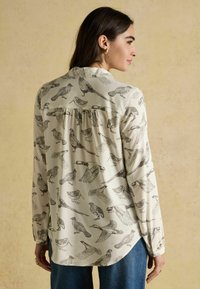 Long-sleeve button-up shirt in cream fabric with bird patterns in dark ink. Relaxed fit, rounded hem, and pleated yoke detail at the back.