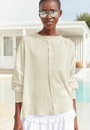 Világos bézs bordázott, hosszú ujjú felső, lekerekített nyakkivágással és oldalsó hasítékokkal. Egy fehér, fodros szoknyával párosítva. Tükörfényes napszemüveget visel.