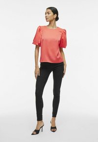 Blusa di satin corallo con maniche a sbuffo, scollatura rotonda, taglio dritto. Abbinata a jeans skinny neri e tacchi aperti neri.