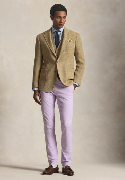 Άνδρας με μπεζ σακάκι, μπλε πουκάμισο με καρό σχέδιο, σκούρο μπλε γραβάτα, κίτρινο μαντηλάκι τσέπης, ροζ παντελόνι και καφέ δερμάτινα μοκασίνια, στέκεται στο απλό φόντο.