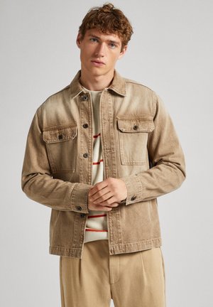 Chaqueta de mezclilla beige con botones en el frente, dos bolsillos en el pecho y un cuello. Lleva puesta una sudadera crema con rayas rojas. Textura suave.