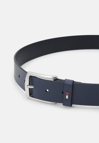 Cintura in pelle blu navy con fibbia in metallo argentato, dotata di un piccolo tag con logo rosso e bianco vicino alla fibbia. Texture liscia, design minimalista.