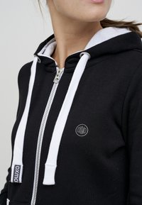Čierna mikina na zips s bielymi šnúrkami, strieborným zipsom a oválnou nášivkou s logom. Obsahuje podloženú kapucňu a mäkkú, textúrovanú látku.