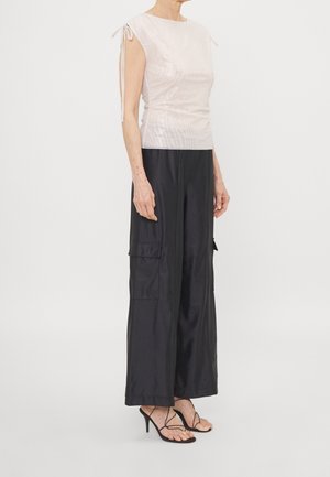 Donna che indossa un top crema senza maniche con dettagli annodati e pantaloni neri a gamba larga con tasche cargo, abbinati a décolleté nere con cinturini.