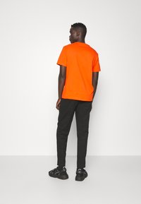 T-shirt arancione in cotone con maniche corte, abbinato a pantaloni neri e scarpe da ginnastica nere. Il design è semplice, senza motivi visibili.