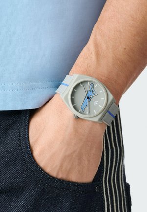 Mano di uomo nella tasca dei jeans scuri che indossa un orologio da polso grigio con un logo script blu sul quadrante grigio chiaro e cinturino in tessuto.