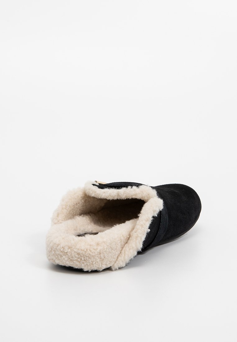 Chausson en suède noir avec doublure en fausse fourrure crème, design ouvert à l'arrière et forme de peau de mouton arrondie. Agrémenté d'un simple détail décoratif en métal.