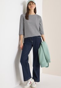 Femme portant une chemise grise rayée, un jean bleu foncé, des baskets blanches et une casquette vert clair, tenant un sweat-shirt vert clair plié.