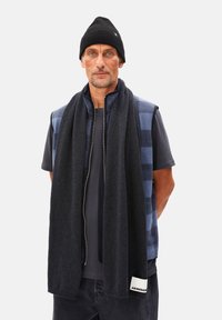 Gilet à carreaux bleu et gris sur un t-shirt noir, associé à une longue écharpe noire. Le bonnet est noir, complétant le look en couches.