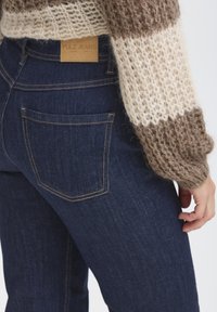 Džínové kalhoty v tmavě modré barvě, s žlutým prošíváním a koženou nášivkou s nápisem "PULZ JEANS." Obsahuje detail zadní kapsy.