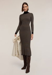 Czarna i beżowa dzianinowa sukienka w paski z wysokim dekoltem, dopasowanym fasonem i rozkloszowanym dołem. Modelka trzyma kremowy płaszcz i ma na sobie buty w wężowy wzór.