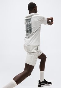 Άνδρας που φοράει ανοιχτό γκρι μπλουζάκι με σχέδιο και αντίστοιχο σορτς τεντώνει τα χέρια ενώ στέκεται στο ένα πόδι, φορώντας μαύρα αθλητικά παπούτσια και λευκές κάλτσες.