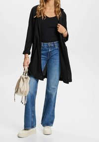 Frau in schwarzem langem Cardigan, schwarzem Top, blauen weit geschnittenen Jeans und weißen Sneakers, hält eine beigefarbene Bucket Bag in der linken Hand.