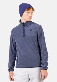 Marinblå fleecepullover med halvdragkedja, hög krage och logotyp på bröstet. Ribbstickad mössa i en mörkare nyans av blått. Slät textur.