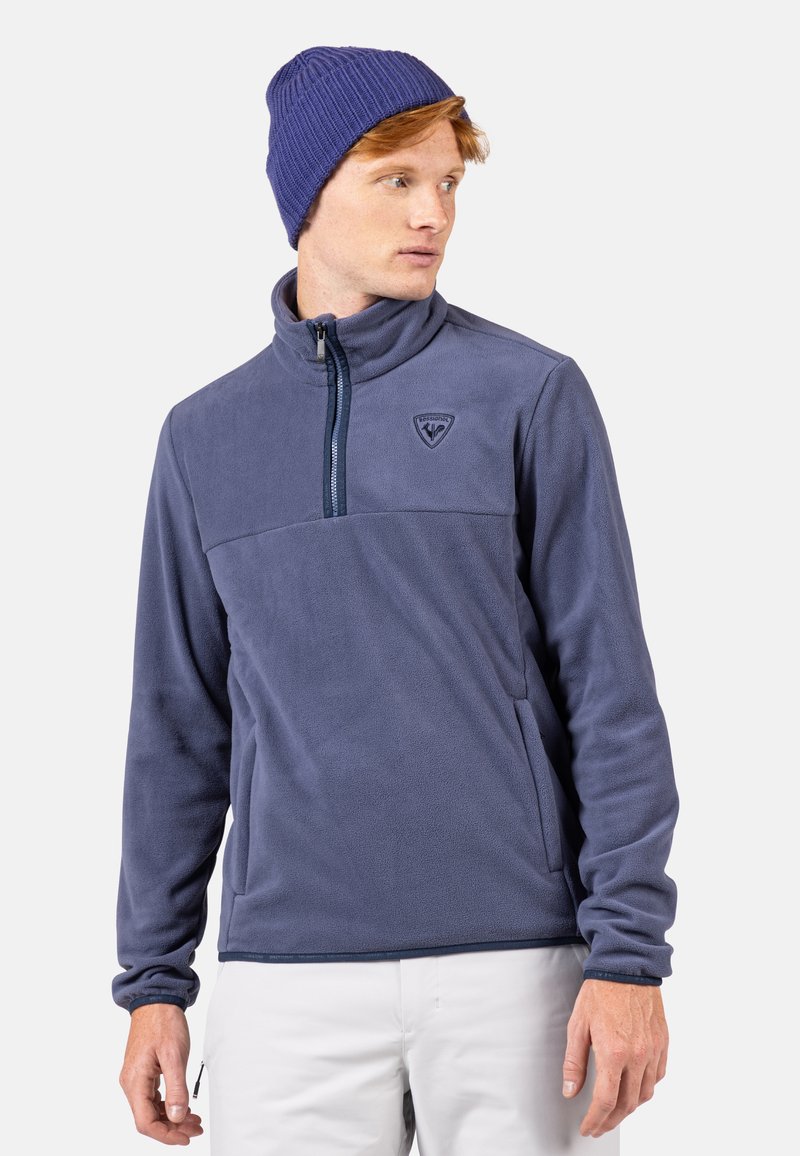 Marinblå fleecepullover med halvdragkedja, hög krage och logotyp på bröstet. Ribbstickad mössa i en mörkare nyans av blått. Slät textur.