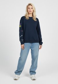 Donna con capelli biondi che indossa un felpa blu navy con stampe colorate sulle maniche, jeans a gamba larga azzurri e sneakers bianche, in piedi rivolta di fronte.