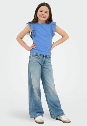 Hymyilevä tyttö seisoo käsivarret lanteilla, pukeutuneena siniseen röyhelöiseen hihattomaan toppiin, leveälahkeisiin vaaleansinisiin farkkuihin ja valkoisiin lenkkareihin vaalealla taustalla.