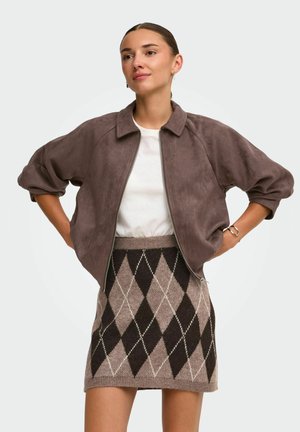 Women wearing brown suede jacket over white shirt and brown argyle-patterned knit skirt, standing with hands on hips.