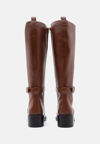Barna bőr, térd fölé érő csizmák sima felülettel, oldalsó cipzárral és díszes csatokkal. Masszív fekete sarkok a stabilitás érdekében.