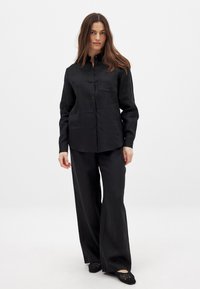 Black linen button-up shirt with a chest pocket, paired with wide-leg black trousers and black crocheted shoes. Smooth texture throughout.
