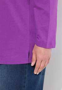 Lila Langarm-T-Shirt mit Seitenschlitzen und glatter Textur, kombiniert mit dunkelblauen Jeans. Nahaufnahme des Saums des Shirts und einer Hand.