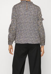 Femme portant un chemisier ample à manches longues à motif floral bleu et beige avec un pantalon noir, vue de dos sur un fond uni.