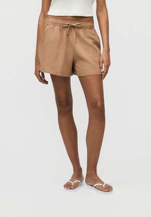 Γυναίκα που φοράει καφέ σορτς με κορδόνι, λευκό crop top και λευκές σαγιονάρες, στέκεται μπροστά σε ένα απλό ανοιχτόχρωμο φόντο.