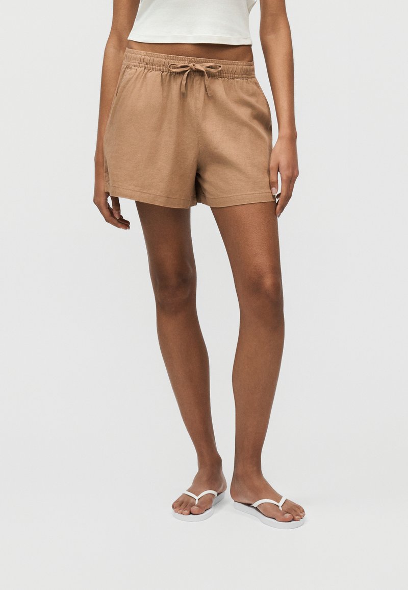 Γυναίκα που φοράει καφέ σορτς με κορδόνι, λευκό crop top και λευκές σαγιονάρες, στέκεται μπροστά σε ένα απλό ανοιχτόχρωμο φόντο.