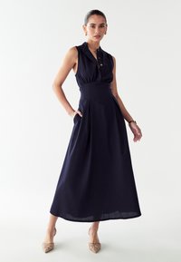 Mornarsko modra maksijska obleka brez rokavov z ovratnikom na gumbe, prilegajočim pasom, stranskimi žepi in zračnim krilom. Gladek, lahek material.