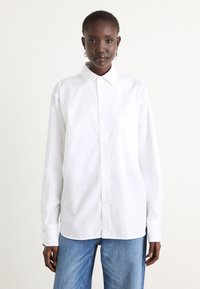 White button-up shirt made of smooth fabric, featuring a pointed collar, long sleeves, and a single chest pocket. Worn with blue jeans.
