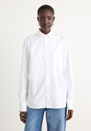 White button-up shirt made of smooth fabric, featuring a pointed collar, long sleeves, and a single chest pocket. Worn with blue jeans.