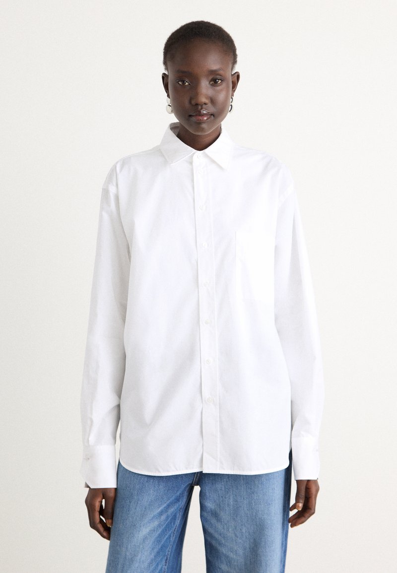 White button-up shirt made of smooth fabric, featuring a pointed collar, long sleeves, and a single chest pocket. Worn with blue jeans.