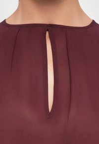 Blusa bordeaux con scollatura a occhio di pernice e chiusura con piccolo bottone, realizzata in tessuto leggero e semi-trasparente con una texture morbida.