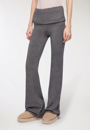 Pantaloni in maglia grigi a campana con una cintura larga e piegata. Tessuto morbido, indossati con pantofole beige.