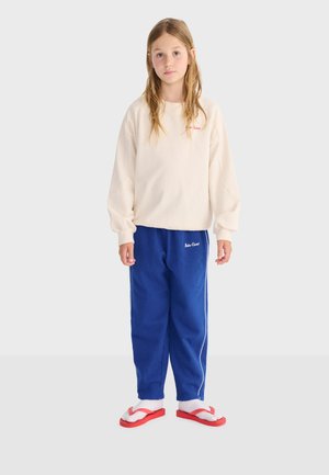 Jeune personne debout portant un sweat-shirt crème, un pantalon de survêtement bleu avec une bande blanche, des chaussettes blanches et des tongs rouges sur un fond uni.