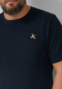 Man die een marineblauw T-shirt draagt met een klein geborduurd vliegend eendje op de borst.