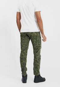 Maskáčové cargo kalhoty v zelených tónech, s bočními kapsami, zúženým střihem a texturou z bavlněné směsi. Kombinovány s bílým jednobarevným tričkem.