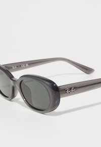Apvalūs pilki permatomi Ray-Ban akinių rėmeliai su tamsiais lęšiais, padėti kampu ant balto fono.