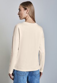 Cremig-beige Langarmshirt mit überschnittenen Schultern, glatter Struktur, rundem Ausschnitt und lockerer Passform, kombiniert mit blauen Jeans.