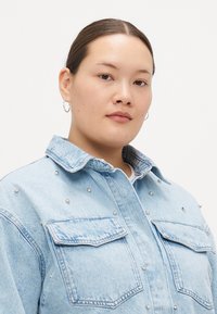 Lys blå denimjakke med sølvnagler, spiss krage og to frontlommer. Jevn tekstur med et avslappet passformdesign.