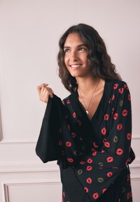 Kvinde med bølget mørkt hår, der smiler, iført en sort kjole med røde og gyldne læbestiftstryk, holder en vid ærme op nær en ensfarvet lys væg.