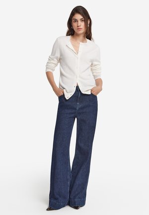 Un gilet blanc à boutons avec des manches longues, porté sur un jean en denim bleu foncé à taille haute et large, créant un look superposé.