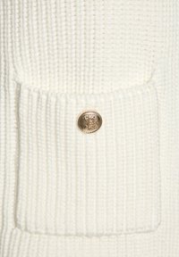 Weißer Strickpullover mit gerippter Textur, ausgestattet mit einer Fronttasche und einem goldenen Knopf mit einem aufwendigen Design.