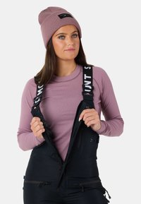 Salopette nera con bretelle regolabili, abbinata a una maglietta a maniche lunghe di un leggero colore viola e un berretto a coste con un patch logo.