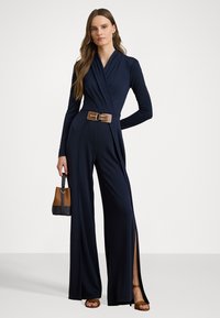 Jumpsuit avvolgente navy con maniche lunghe, cintura strutturata, spacchi laterali e texture liscia. Abbinato a una borsa marrone e nera e sandali.