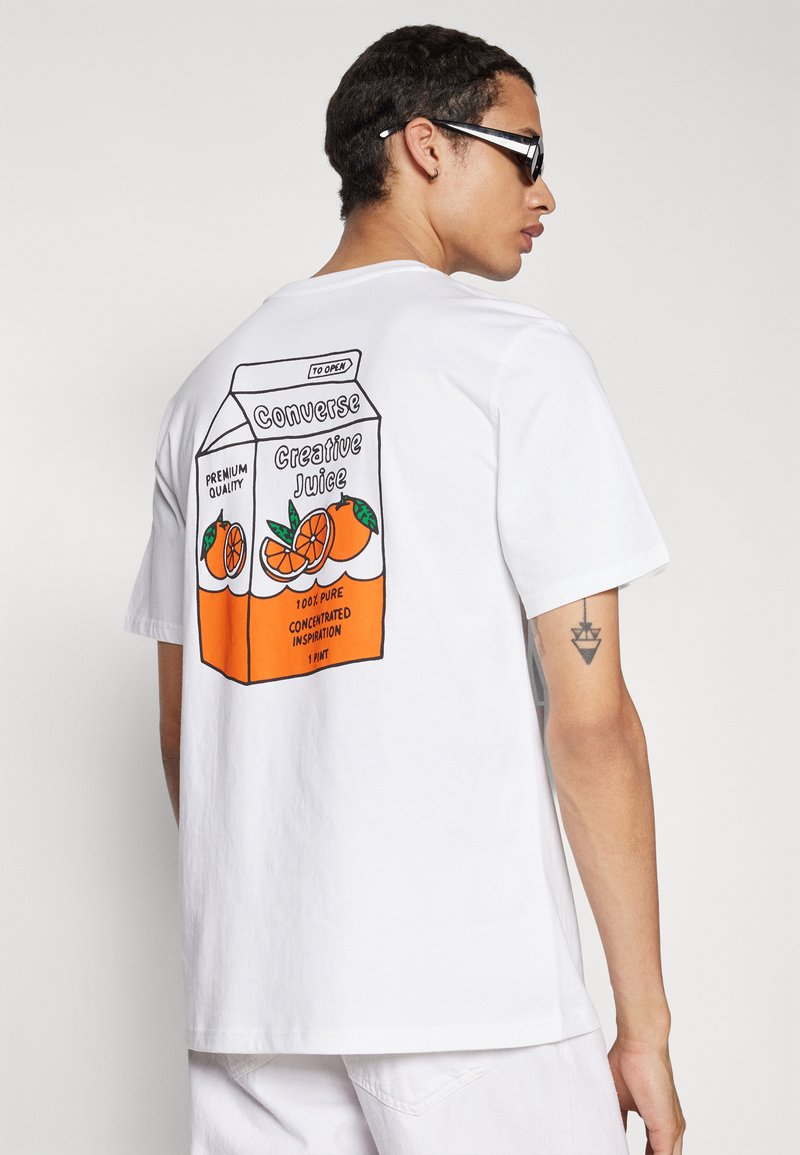 Camiseta blanca de manga corta con un gráfico colorido de un cartón de jugo en estilo de dibujos animados en la parte de atrás, con acentos en naranja y verde.
