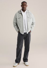 Lichtgrijs jack met rits en capuchon, met twee borstzakken, gedragen over een wit overhemd met knopen en donkere spijkerbroek. Witte sneakers met een beige zool.